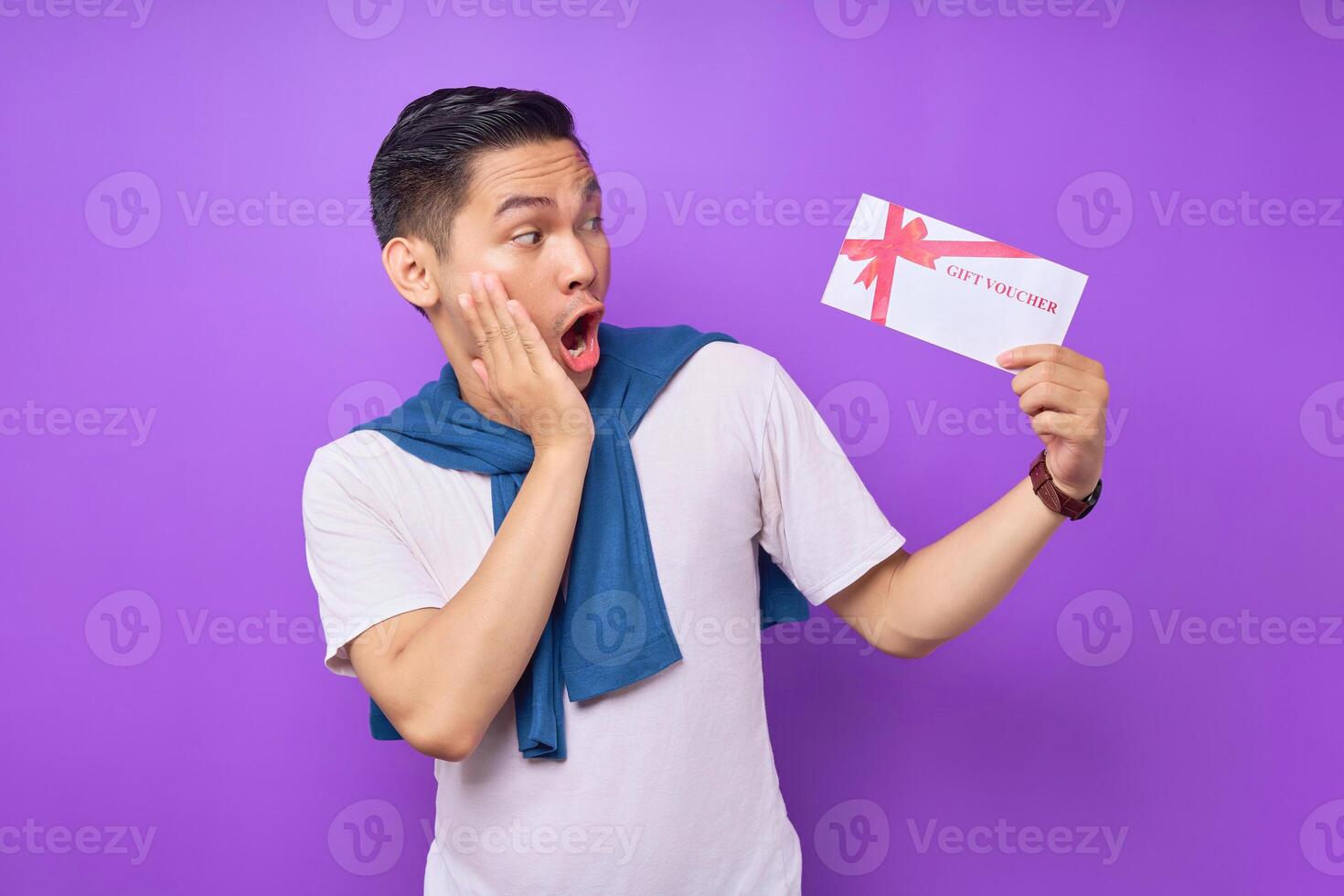 Surprised handsome Asian ethnicity young man 20s wearing casual clothes holding gift certificate coupon voucher card for the store isolated on purple background. People lifestyle concept photo