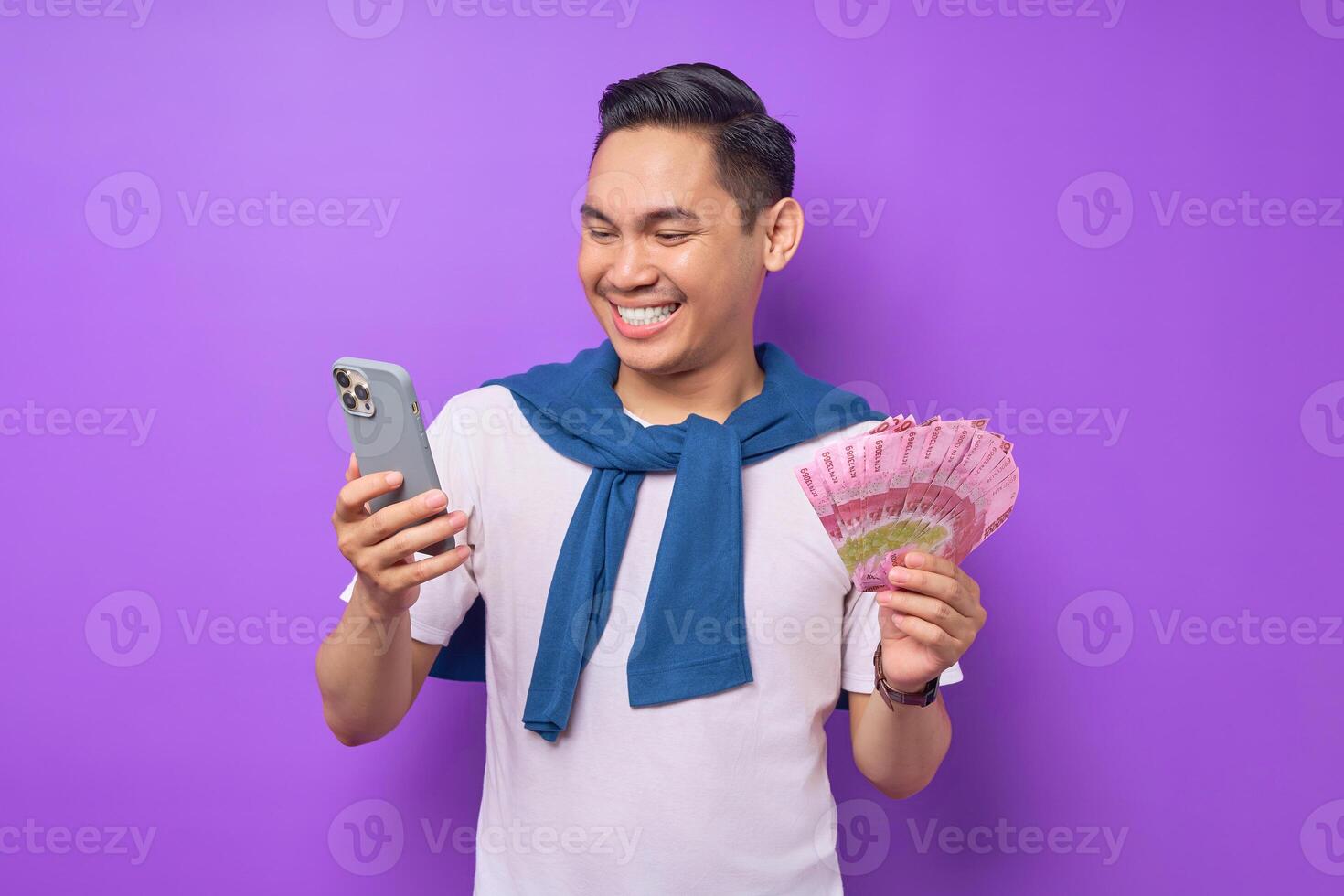 Portrait of smiling young Asian man using mobile phone and holding money banknotes isolated over purple background photo