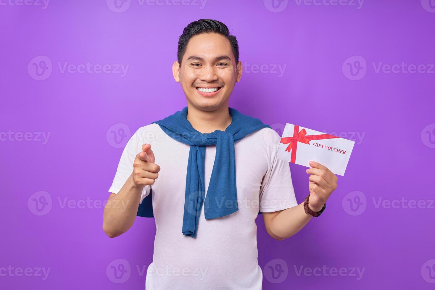 Smiling young Asian man in casual clothes holding gift certificate and pointing finger the camera isolated on purple studio background photo
