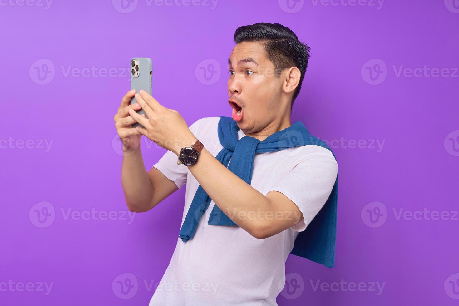 Surprised young Asian man looking at smartphone screen and emotionally reacting to online news isolated over purple background photo