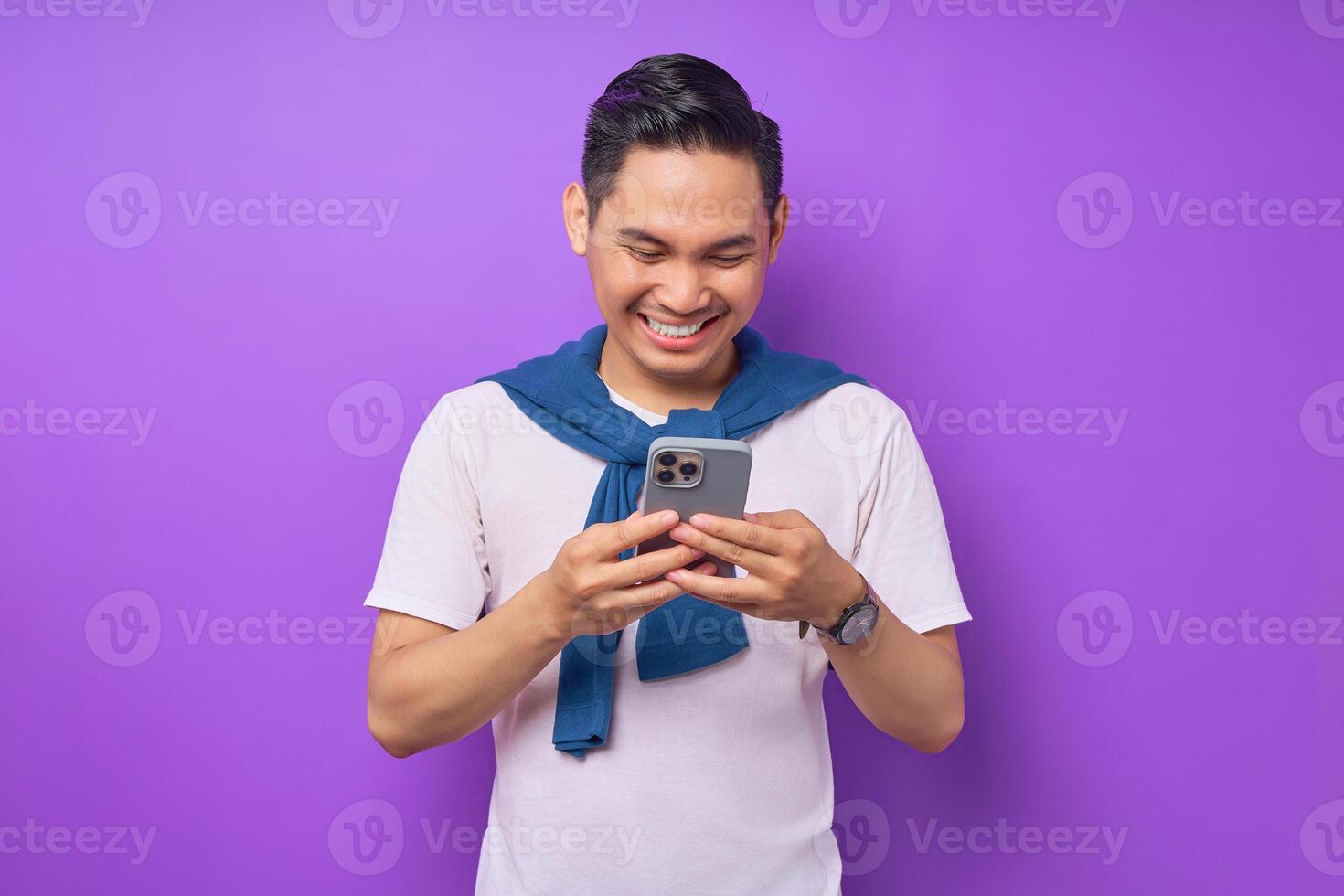 Cheerful young Asian man using smartphone for messaging with friends isolated over purple background photo