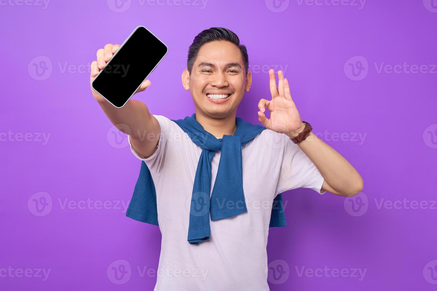 Cheerful young Asian man holding smartphone with blank screen and gesturing okay sign isolated on purple background. Advertising new mobile app, mockup concept photo