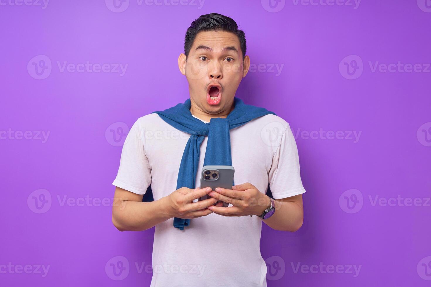 Surpised young Asian man holding smartphone and smiling at camera isolated over purple background photo