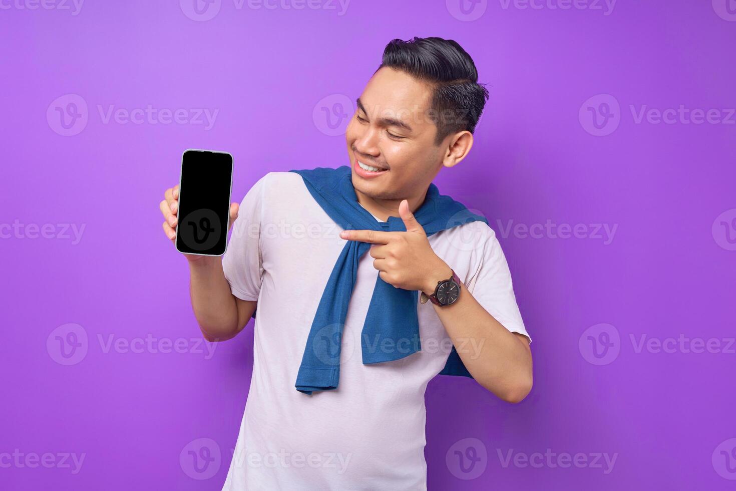 Cheerful young Asian man pointing index finger at empty smartphone screen isolated on purple background. Advertising new mobile app, mockup concept photo
