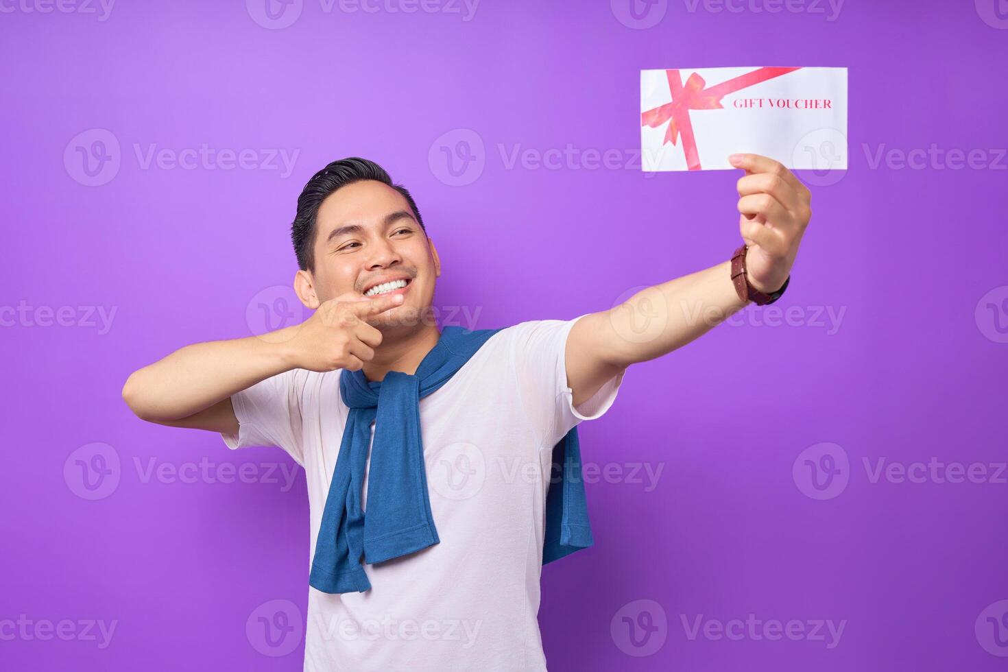 Smiling young Asian man in casual clothes pointing a finger at gift voucher certificate and looking at the camera isolated on purple studio background photo