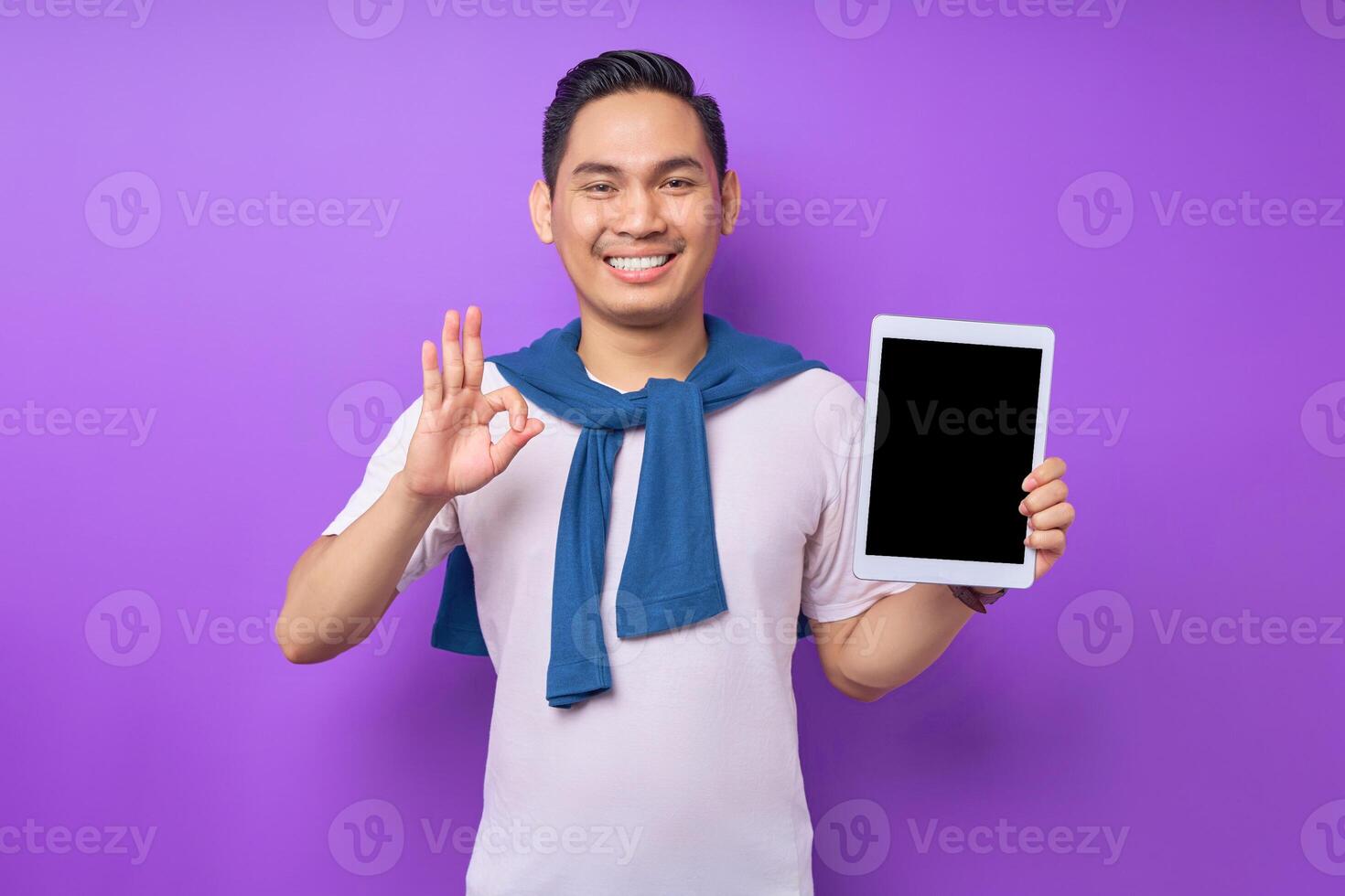 Cheerful young Asian man in casual clothes showing digital tablet with empty screen for mockup and okay sign isolated over purple background photo