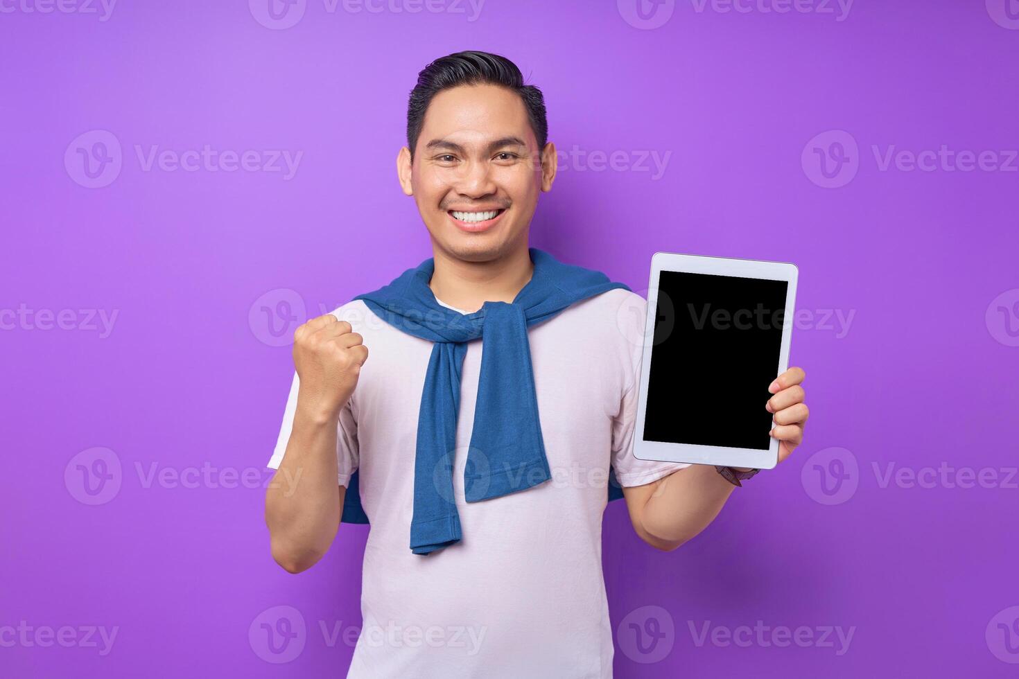 Excited young Asian man in casual clothes showing digital tablet with empty screen for mockup and celebrating success isolated over purple background photo