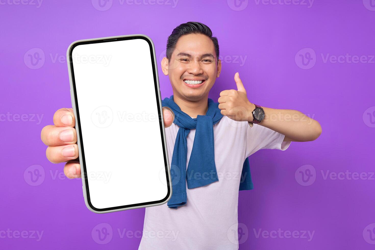 Cheerful young Asian man demonstrating smartphone with empty screen, showing thumb up gesture isolated on purple background. Advertising new mobile app, mockup concept photo