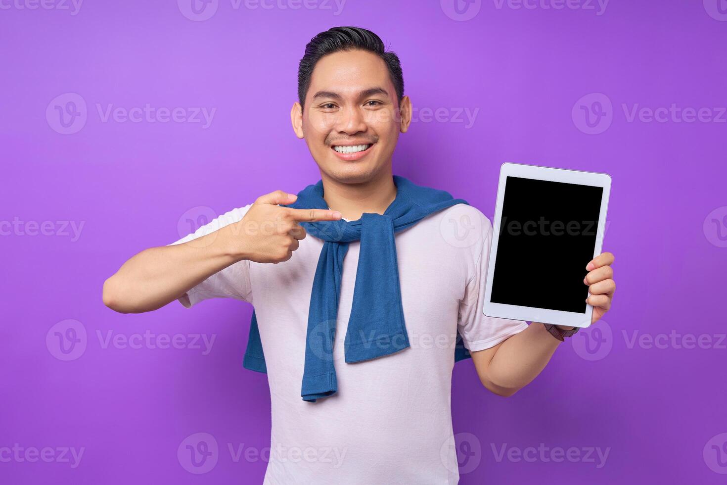 Smiling young Asian man in casual clothes pointing finger at digital tablet with empty screen for mockup isolated over purple background photo