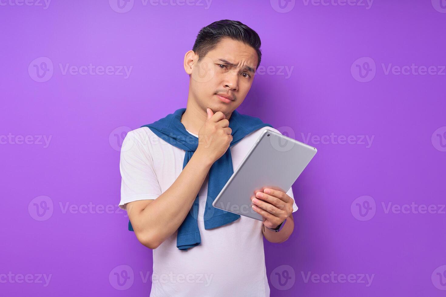 Portrait of pensive young Asian man in white t-shirt touching chin and holding digital tablet isolated on purple background photo