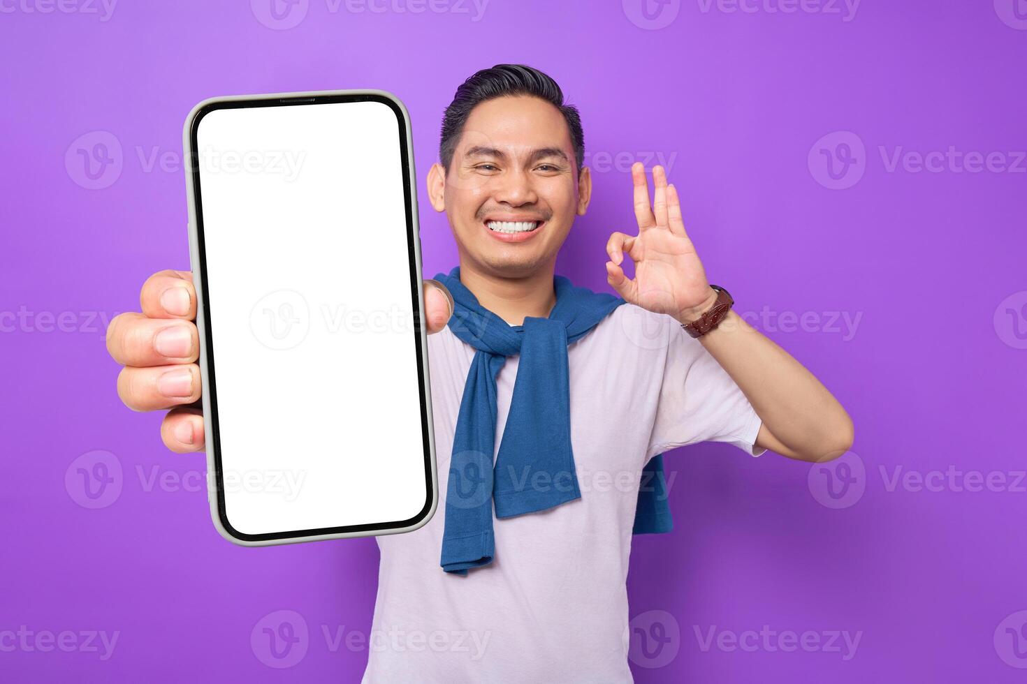 Cheerful young Asian man holding smartphone with blank screen and gesturing okay sign isolated on purple background. Advertising new mobile app, mockup concept photo