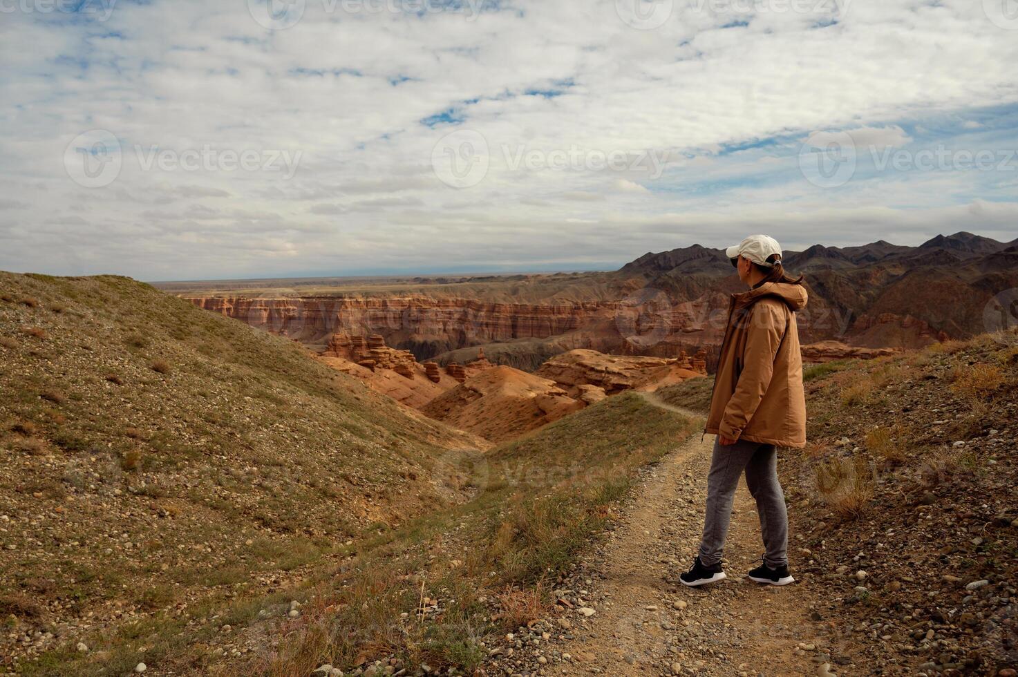 a woman in a brown coat and hat is standing on a dirt road photo