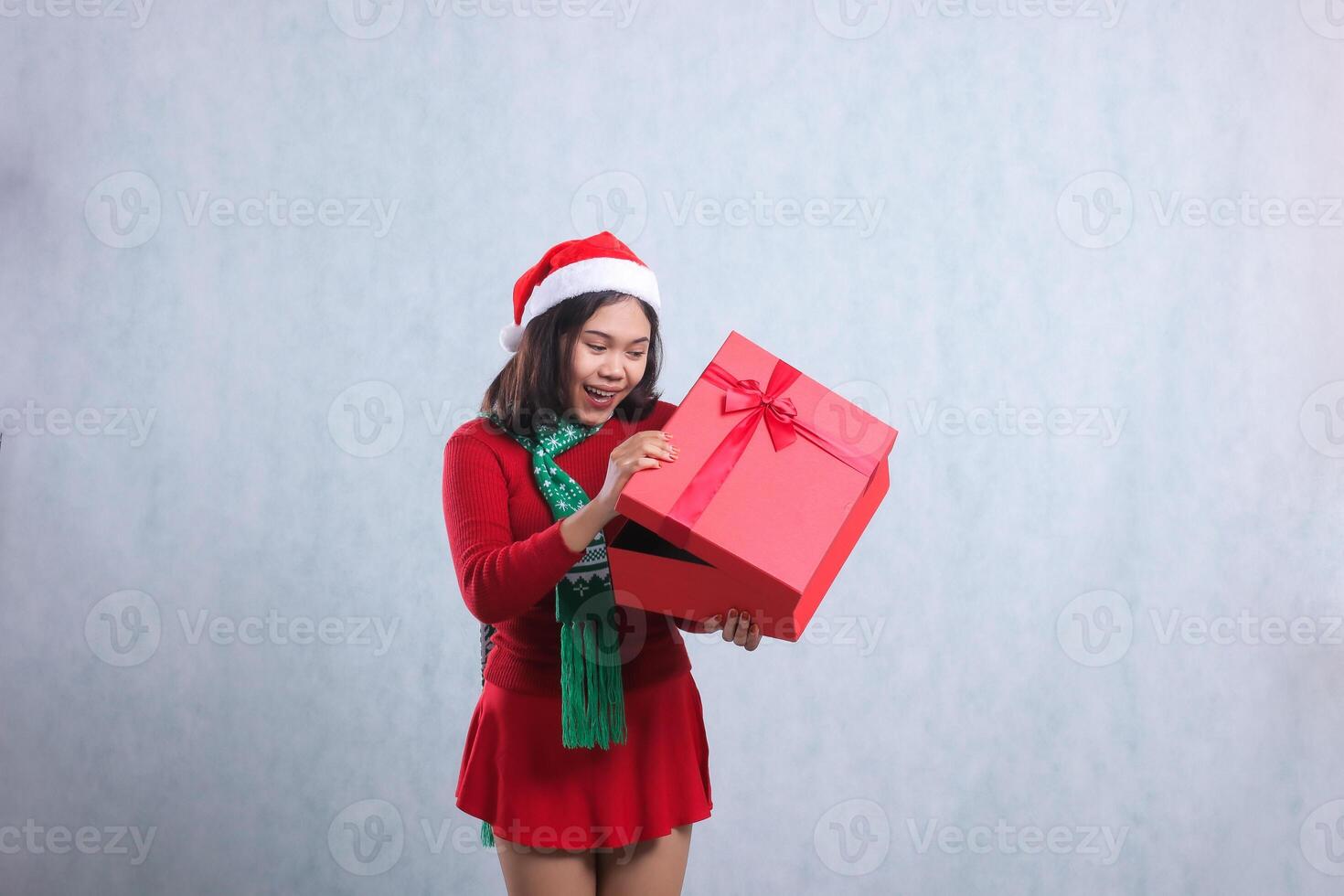 expression of female wearing christmas sweater, santa hat and scarf, happy looking at the contents of the box both hands peeking at the contents of merry christmas box, isolated on white background photo