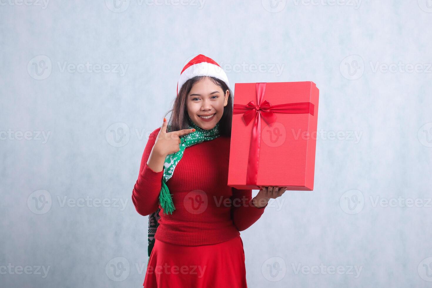 expression of young asian woman wearing christmas sweater, santa hat and scarf, happy looking at camera hand carrying red merry christmas box gift while pointing at it, isolated on white background photo