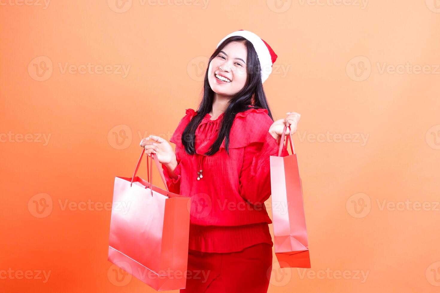 Asian young girl expression wearing red blouse and santa hat, standing right sideways happily to the front both hands lifting red bag of christmas gifts, isolated on orange background photo