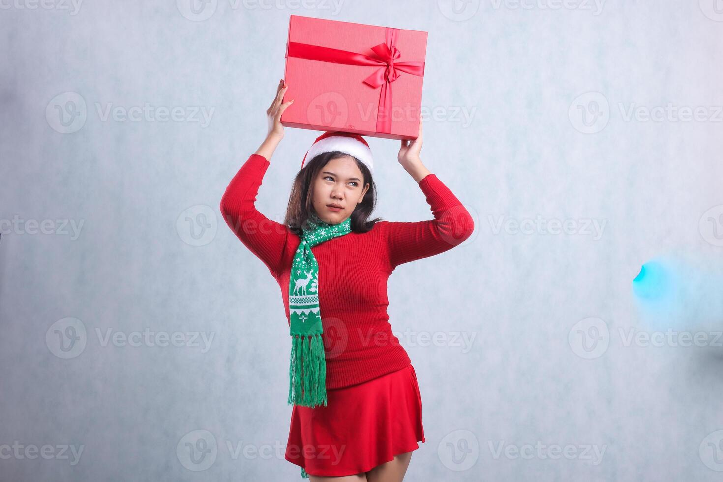 expression of teenage woman wearing christmas sweater, santa hat and scarf, pouting look left both hands raised up red merry christmas gift box, isolated on white background photo