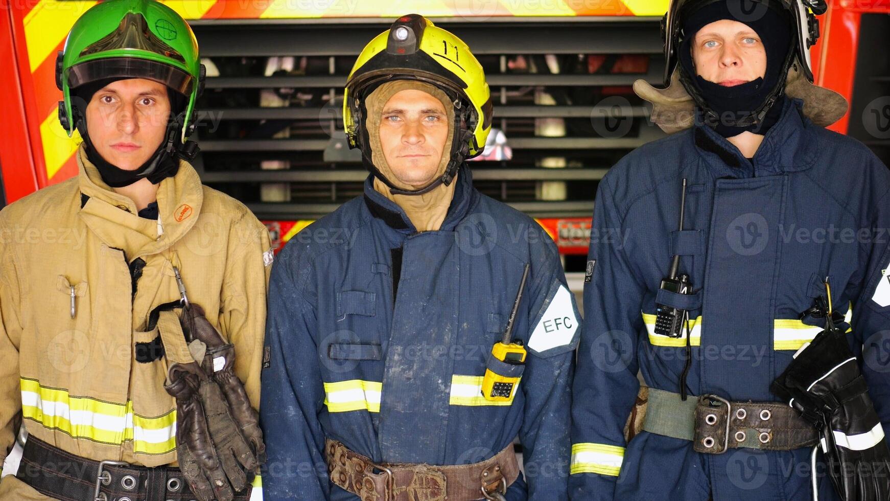 Portrait of male firefighters in helmets and protective uniforms standing near a big red car. Young confident firemen in full equipment posing against the background of a fire engine at station photo