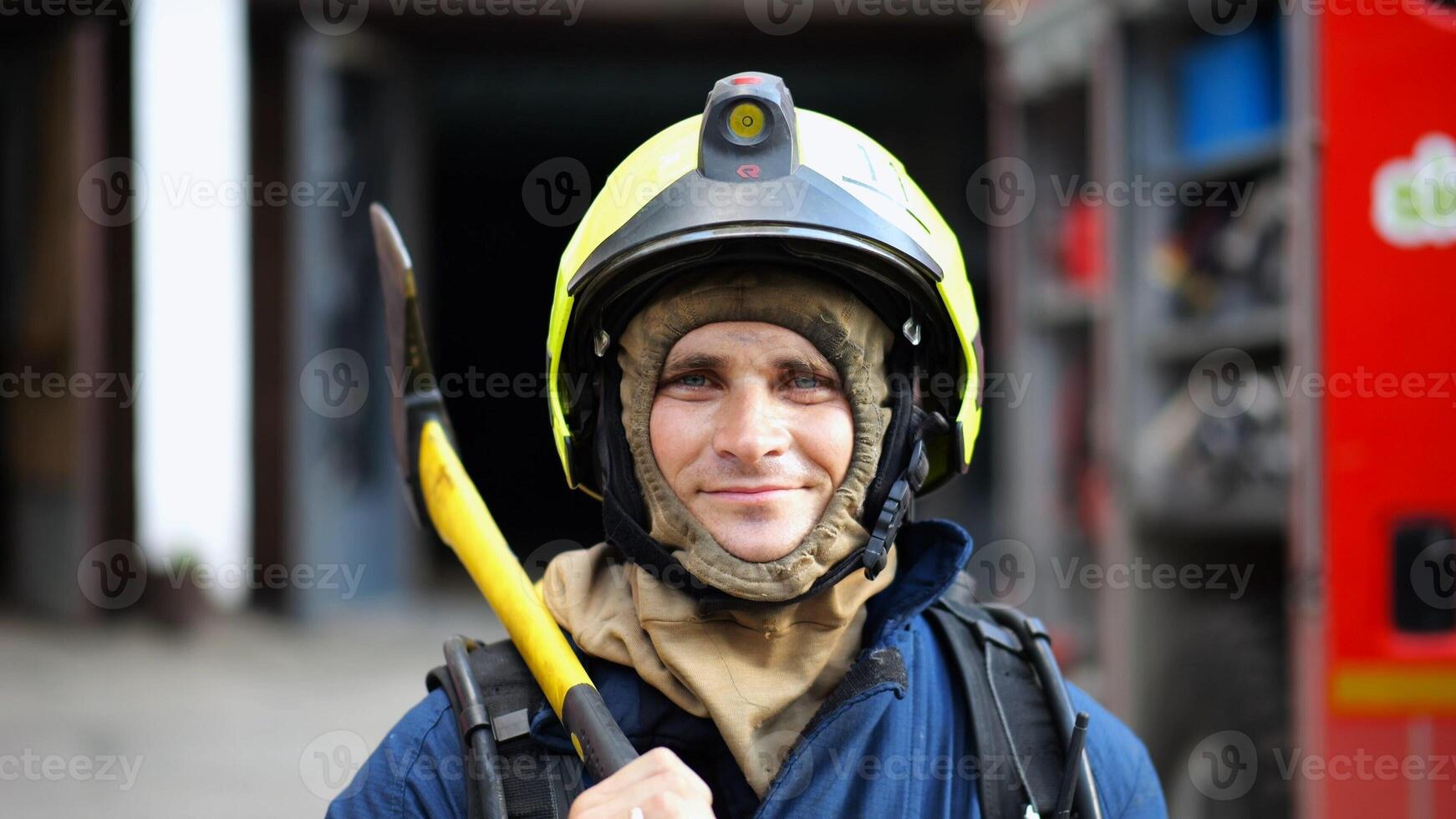 Portrait of happy male fireguard with axe standing near a fire engine. Young smiling fireman looking at camera in full equipment after fire near big red truck. Saving lives and heroic profession photo