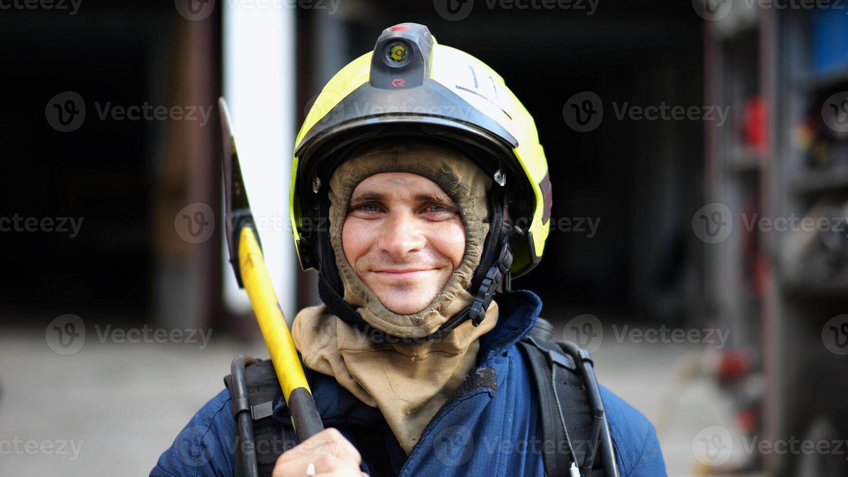 Portrait of happy male fireguard with axe standing near a fire engine. Young smiling fireman looking at camera in full equipment after fire near big red truck. Saving lives and heroic profession photo