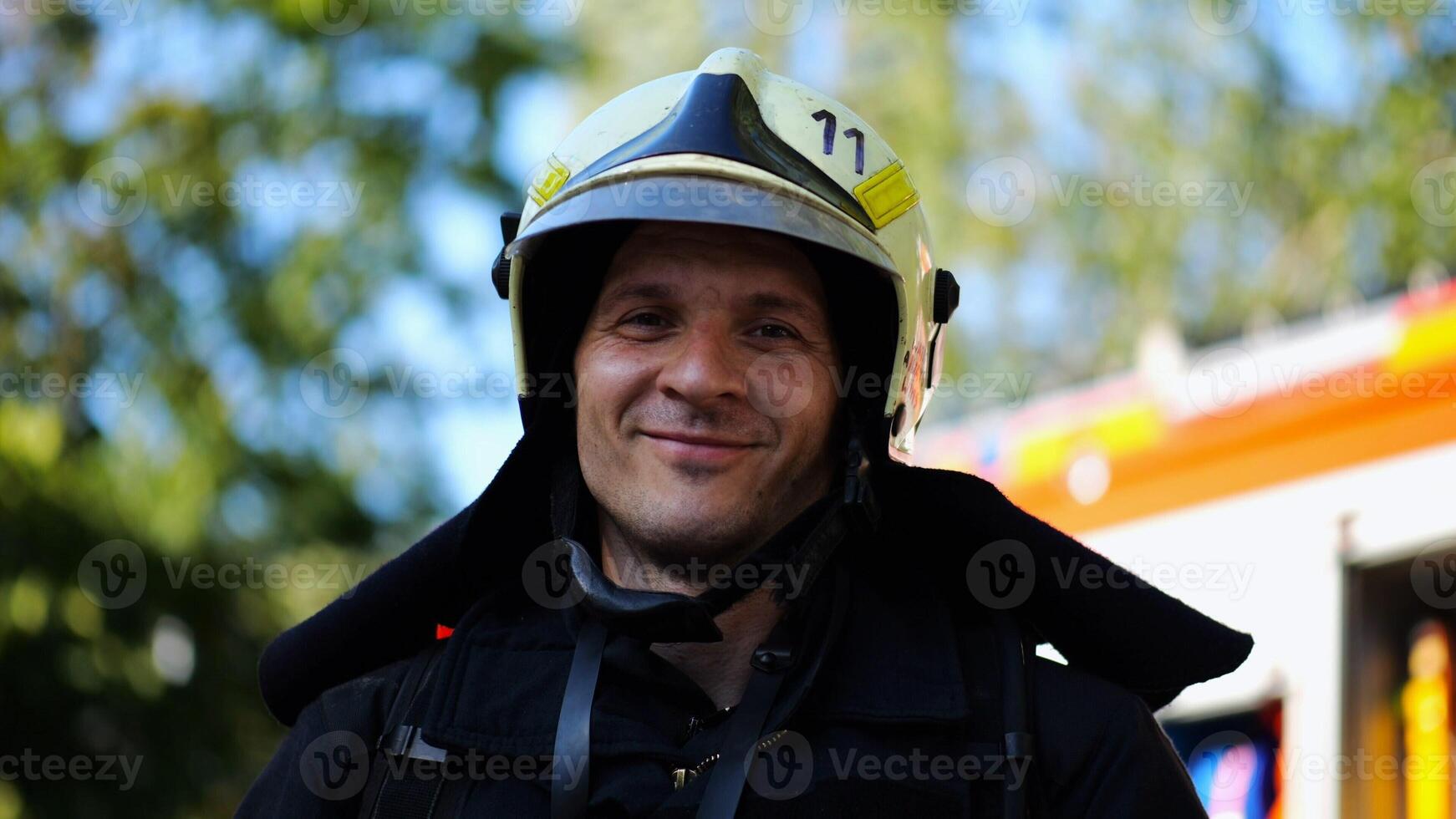 Portrait of young confident fireguard in protective uniform against background of a fire truck. Male firefighter in full equipment looking into camera with positive emotions near big red car. Close up photo