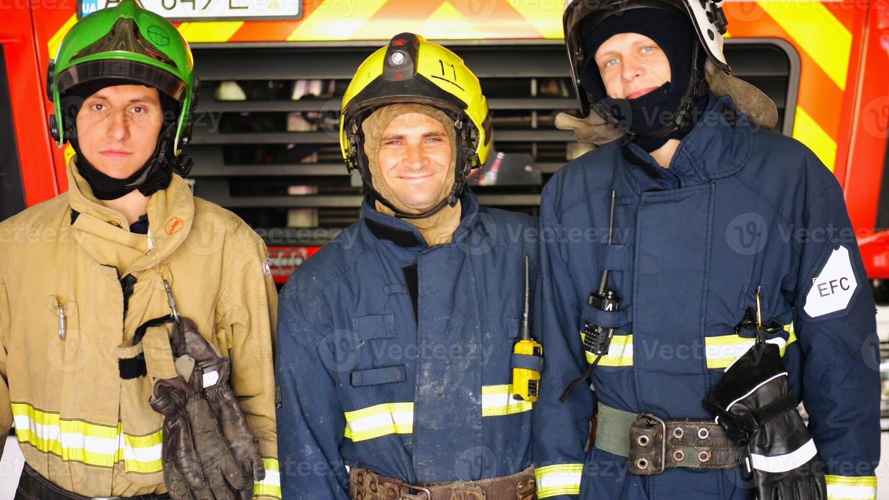 Portrait of young happy fireguards in protective uniforms against background of fire truck at station. Male firefighters in full equipment looking into camera with positive emotions near big red car photo