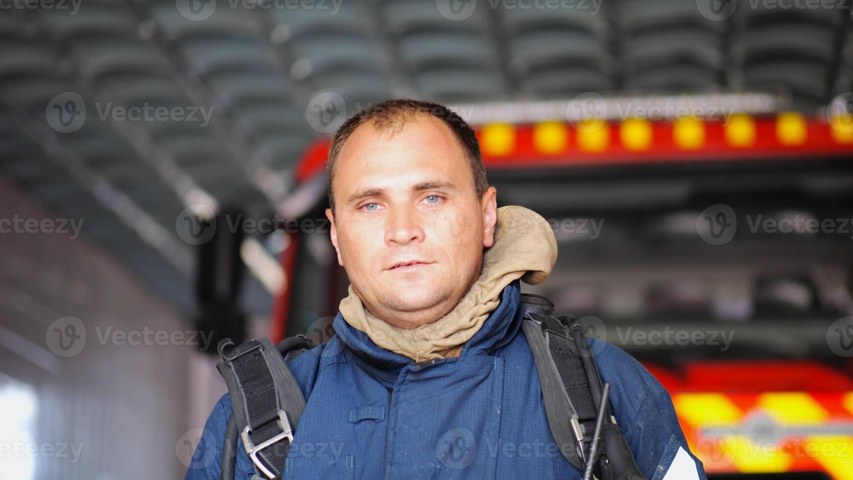 Portrait of tired male firefighter standing near a fire engine at department. Young exhausted fireman in protective uniform looking at camera after fire against the background of big red truck photo