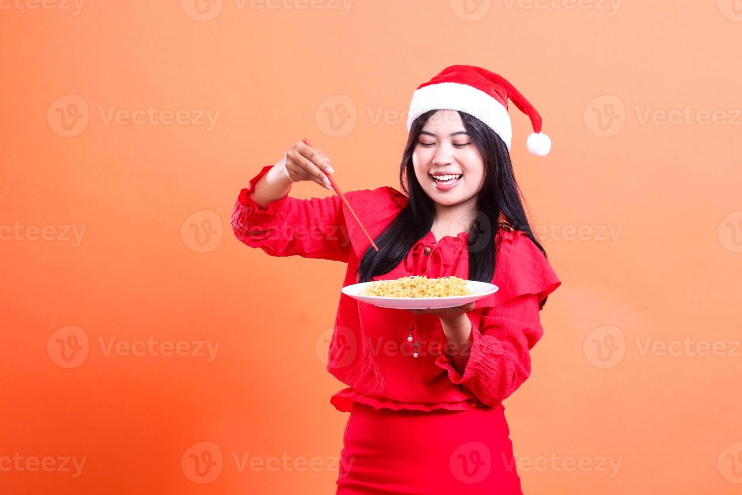 gesture of beautiful women wearing red christmas blouse, Santa hat, cheerful to noodles, hand holding chopsticks towards noodles and white plate filled with noodles, isolated on orange background photo