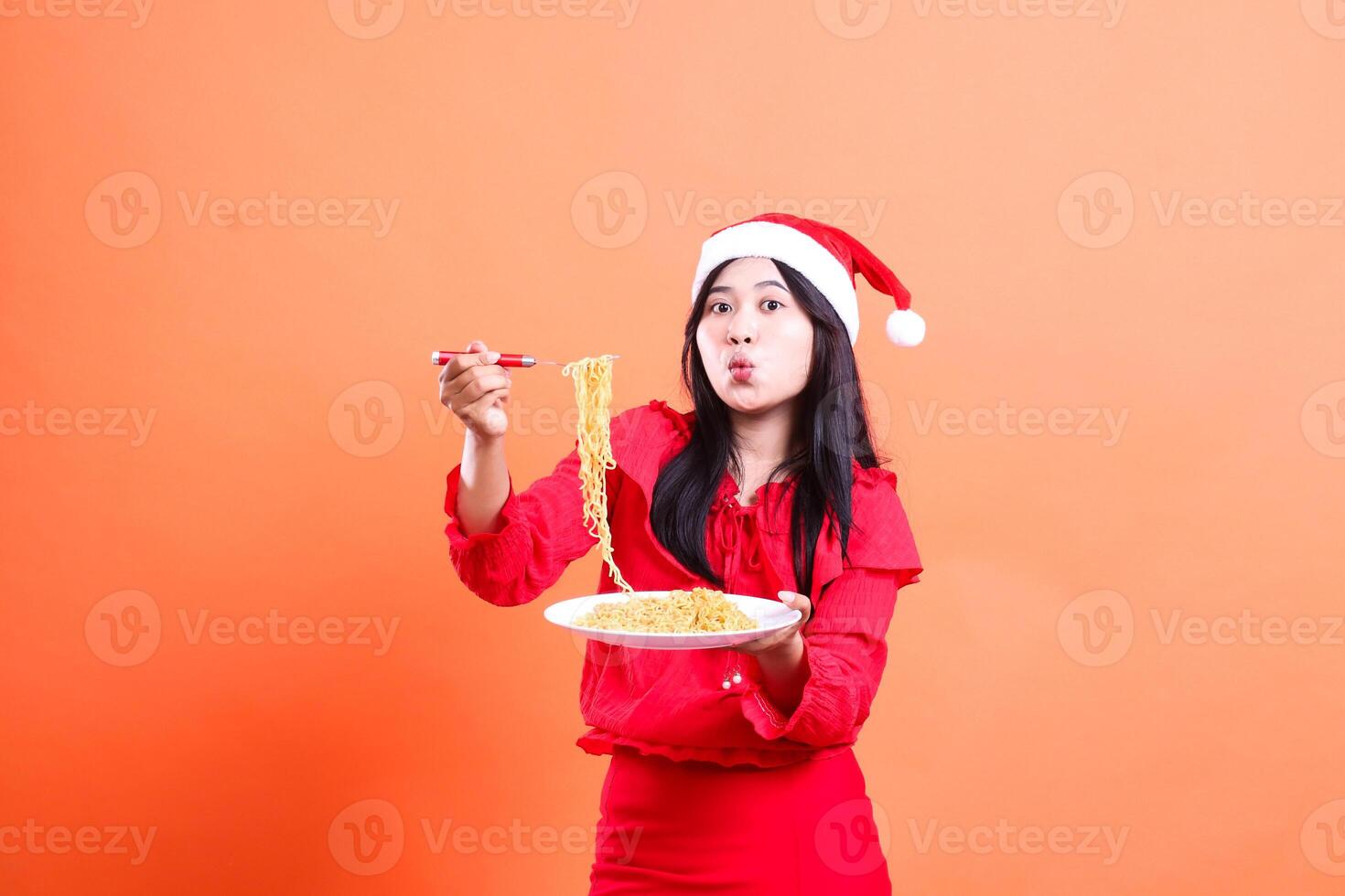 portrait of Asian women wearing Christmas blouse, Santa hat, amazed showing noodles front view, hand holding noodle fork and white plate filled with noodles front view, isolated on orange background photo