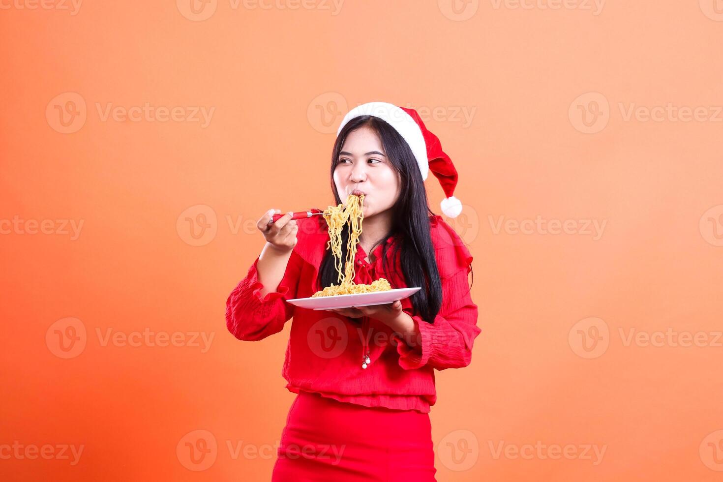 expression of young girl wearing christmas dress, santa hat, enjoy noodles look right, hand holding fork filled with noodles from white plate, isolated on orange background photo