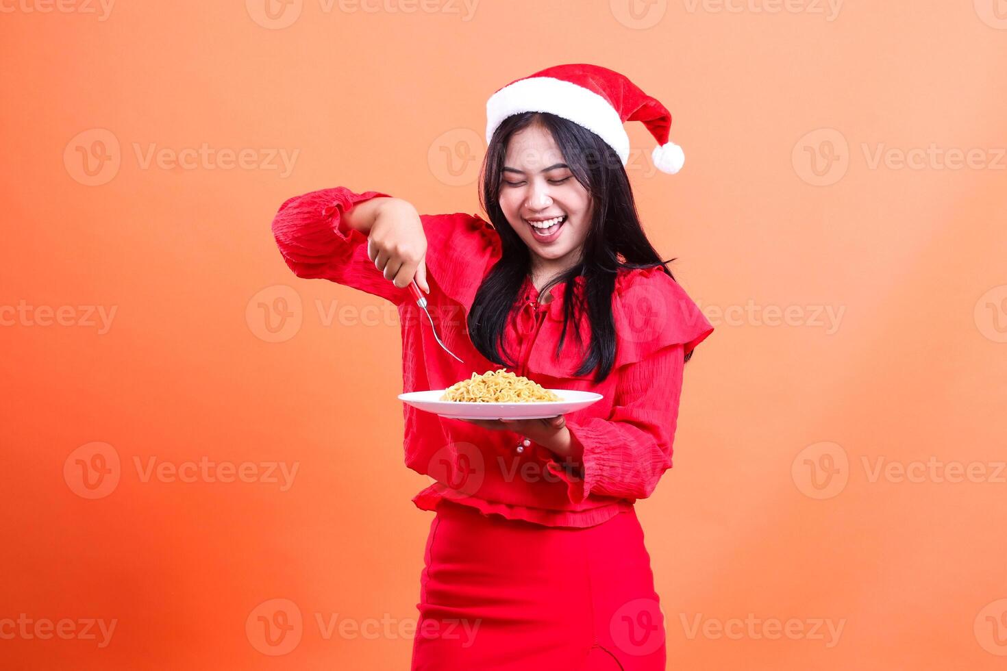 gesture of beautiful female wearing red christmas blouse, Santa hat, cheerful to noodles, hand holding fork about to stab noodles in white plate celebrating christmas, isolated on orange background photo