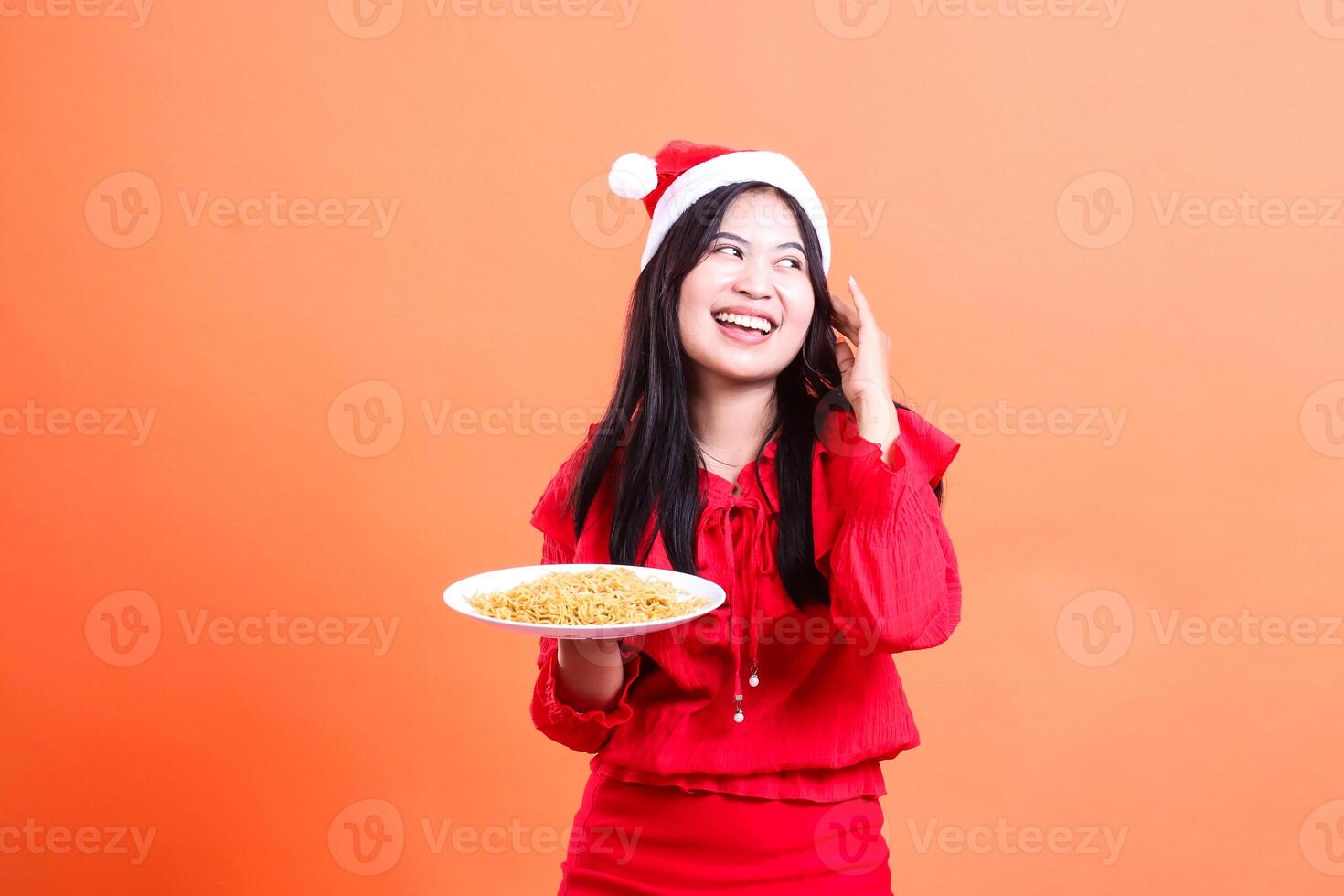 portrait of a ladies wearing Christmas clothes, Santa hat, cheerful to the left, hands holding a white plate filled with noodles to the right and touching her long hair, isolated on orange background photo