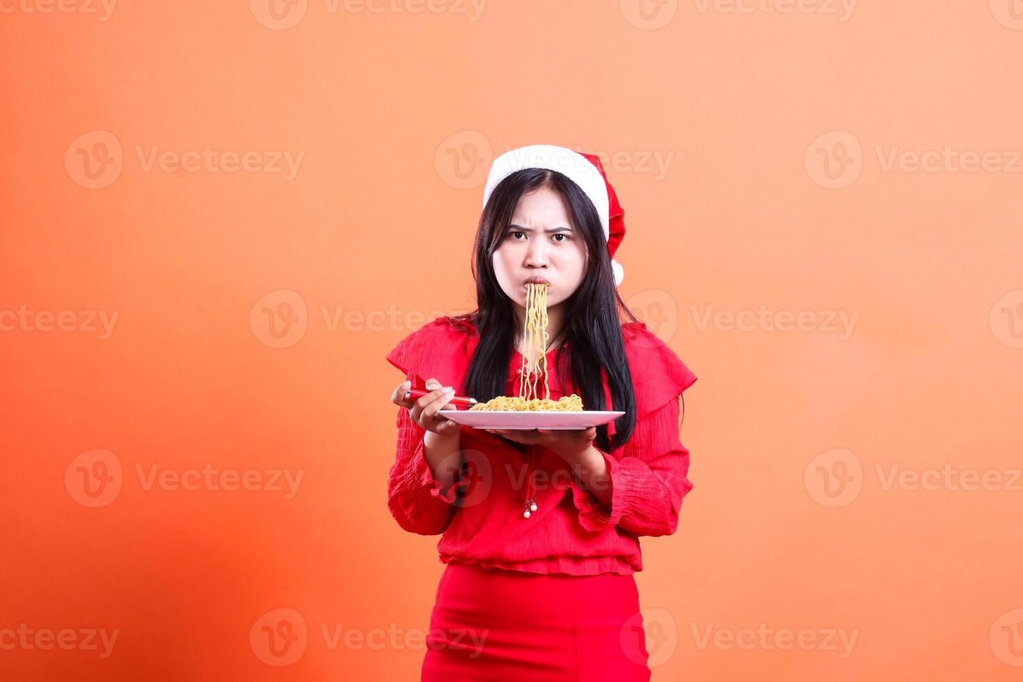 expression of adult female wearing christmas dress, santa hat, pouting slurping noodles front view, hands holding fork and white plate filled with noodles, isolated on orange background photo