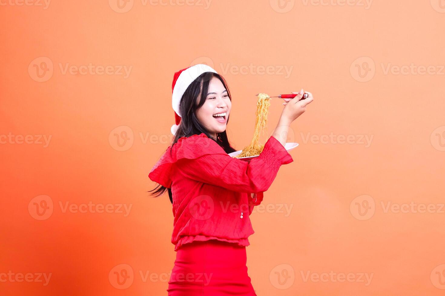 gesture of beautiful woman wearing red christmas blouse, Santa hat, cheerful left slant looking at noodles, hand holding noodle fork and white plate filled with noodles, isolated on orange background photo
