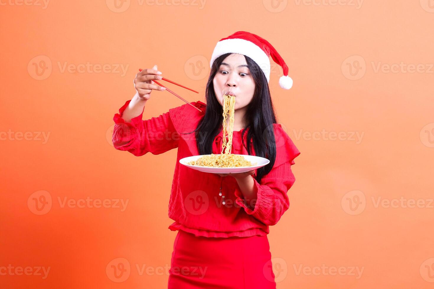 indonesia woman beauty shocked slurping and looking at noodles, hands holding chopsticks and white plate filled with noodles, wearing red christmas blouse, isolated on orange background photo