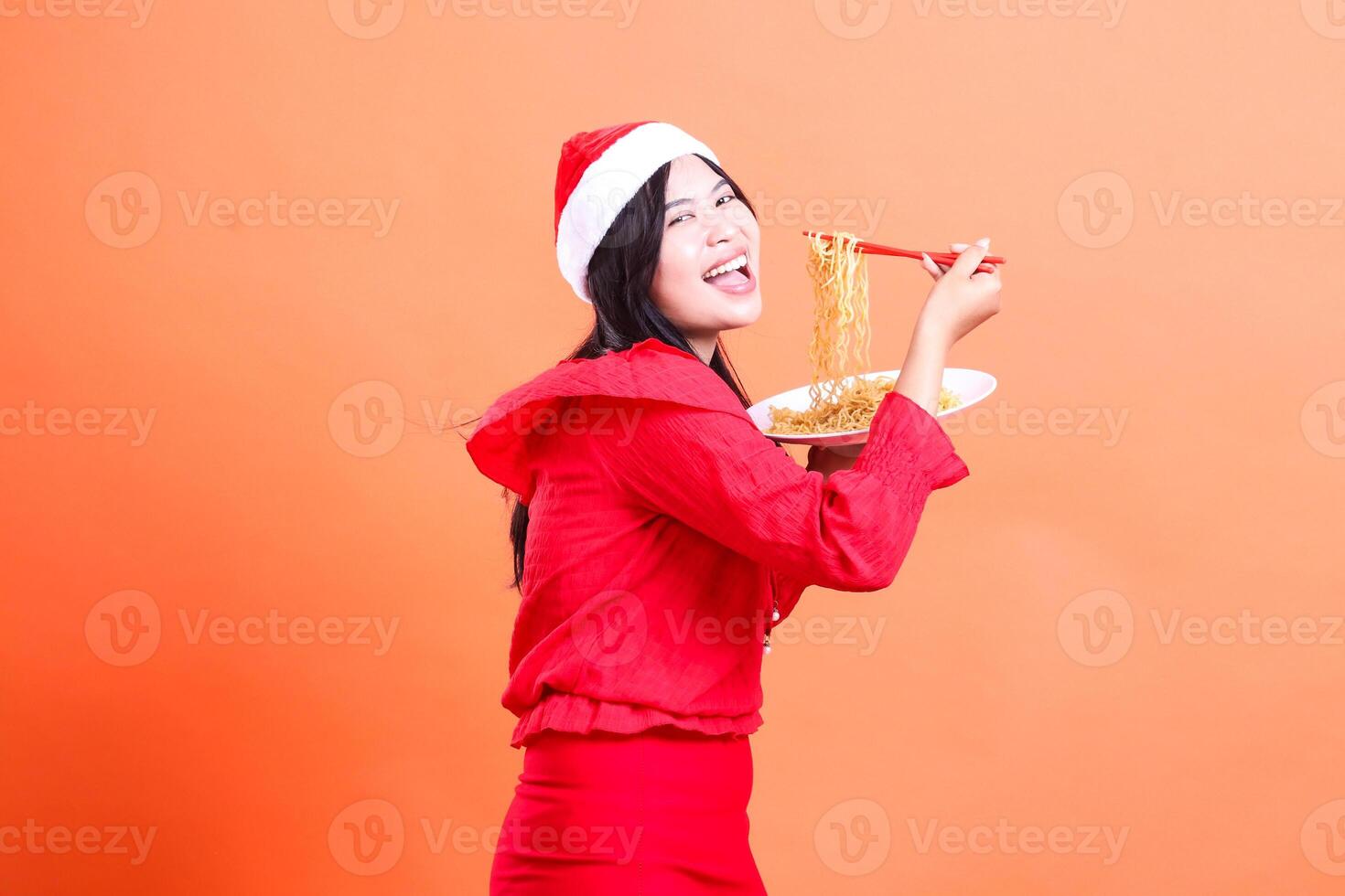 gesture of beautiful woman wearing red christmas blouse, Santa hat, left side laughing to camera, hand holding chopstick to mouth and white plate filled with noodles, isolated on orange background photo