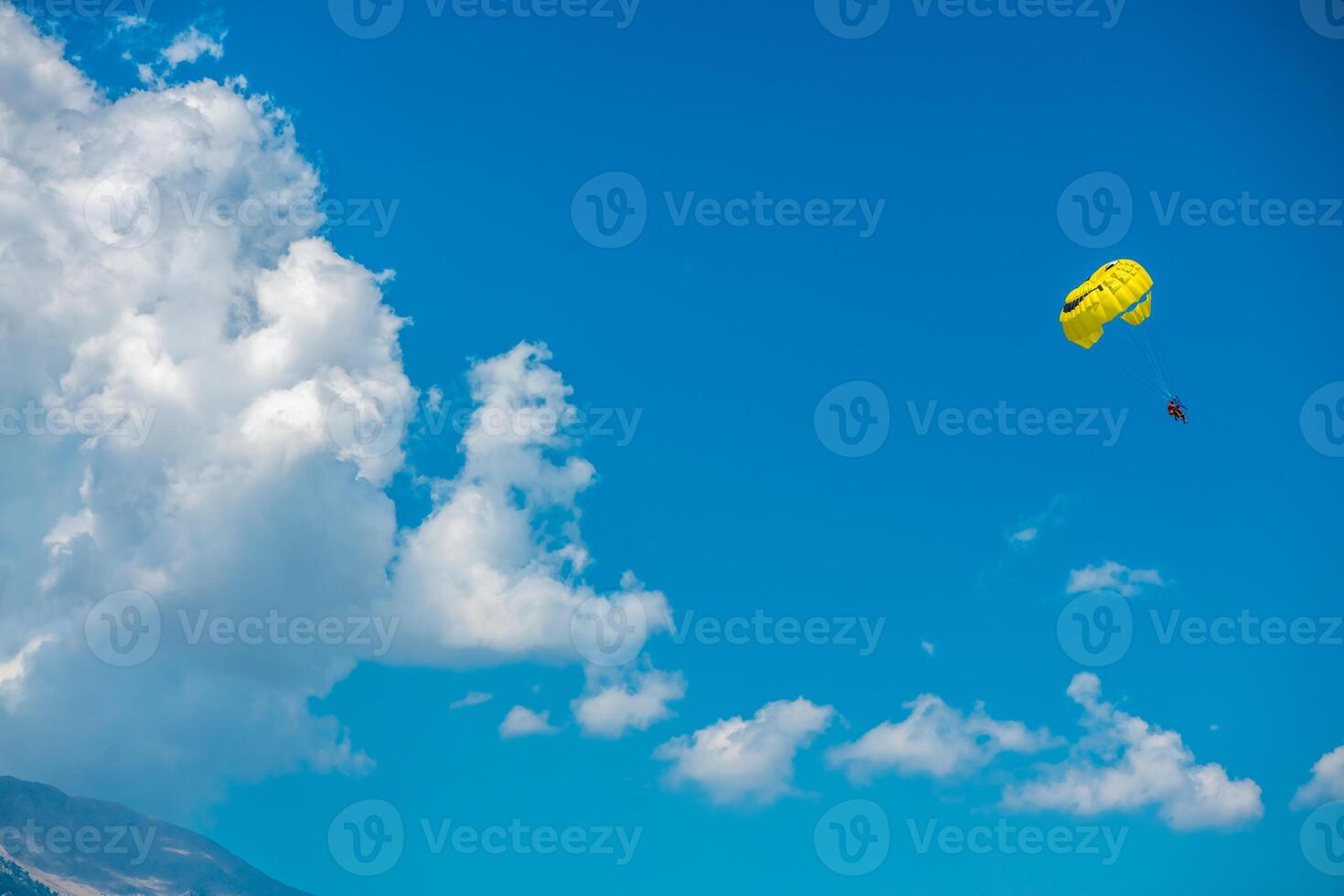 Two people parasailing on a yellow parachute flying against the background of mountains, blue sky and clouds photo