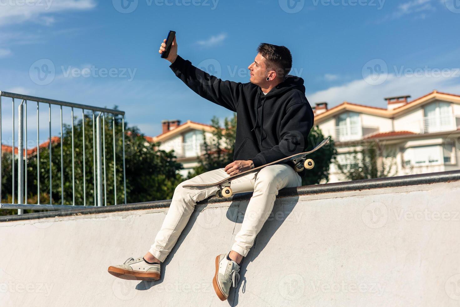 Skater taking a selfie with smartphone while sitting on half-pipe edge in skate park photo
