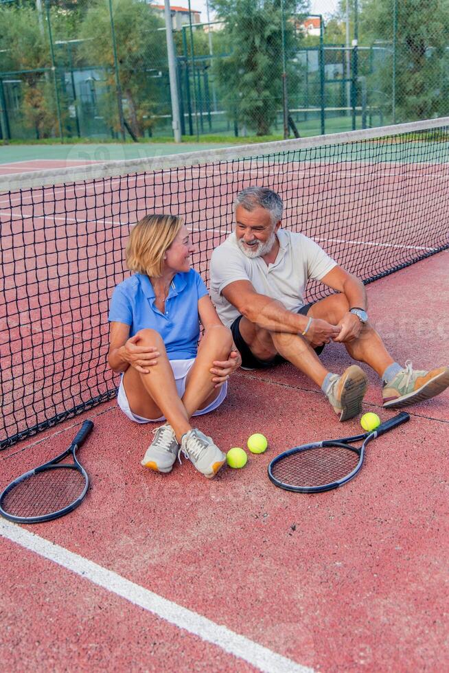 vertical senior tennis couple seated by net with confident expressions and gear photo
