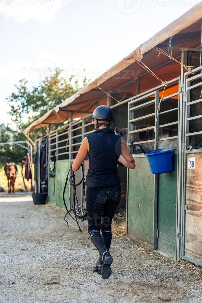 vertical Female rider with a helmet walking through the stables carrying saddles at the ranch photo