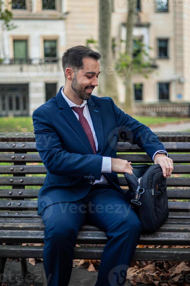 vertical smiling businessman works outdoors, taking a break at a park bench while using his laptop photo