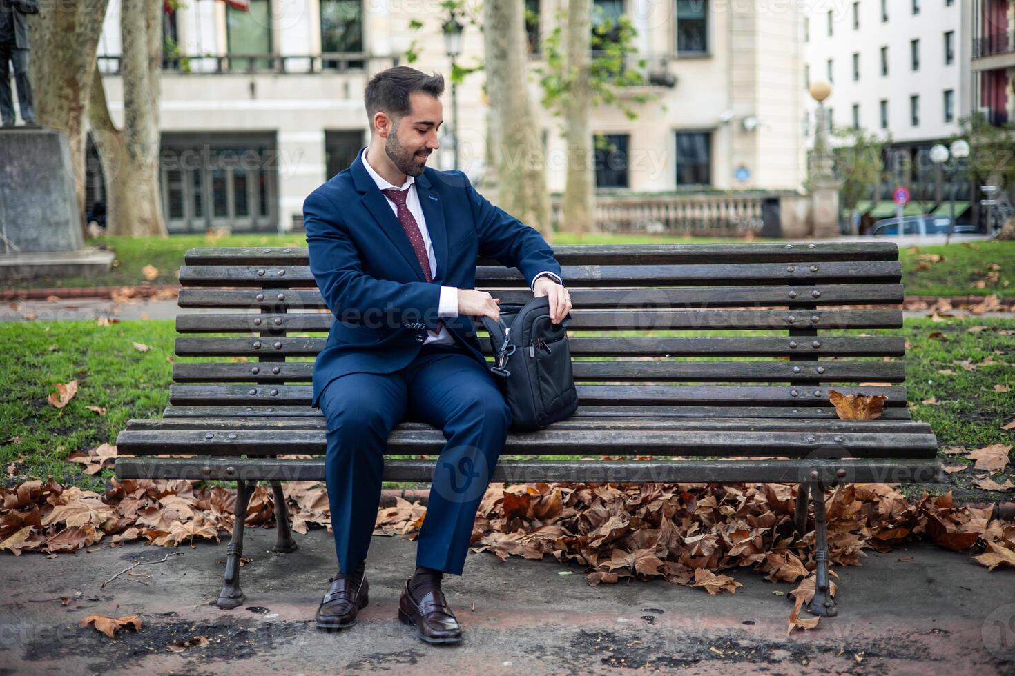 Smiling businessman sitting on a park bench takes his laptop out of the briefcase to work outdoors photo