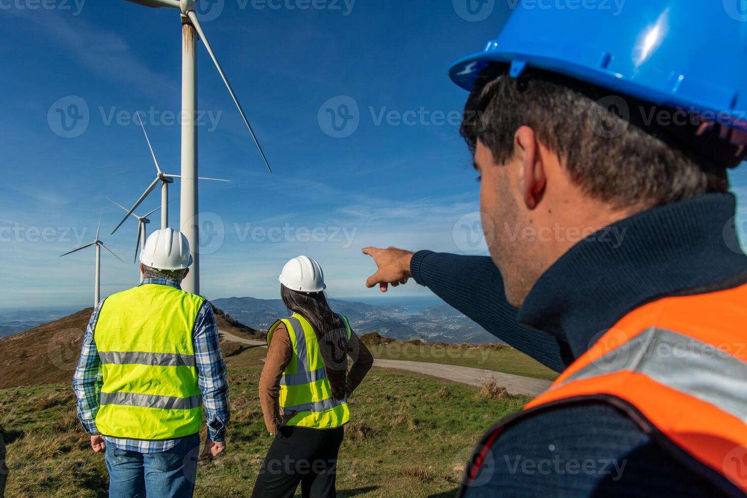 Three multiethnic Engineers pointing at landscape near wind turbines on mountain top photo