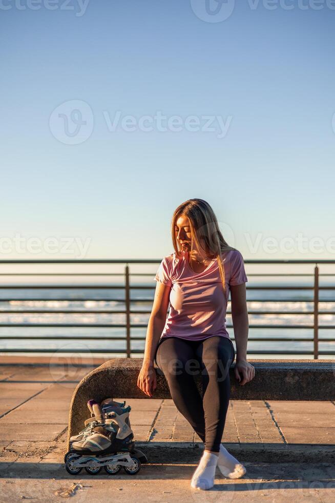 Young sportswoman resting on bench after inline skating by the sea at sunset copyspace photo