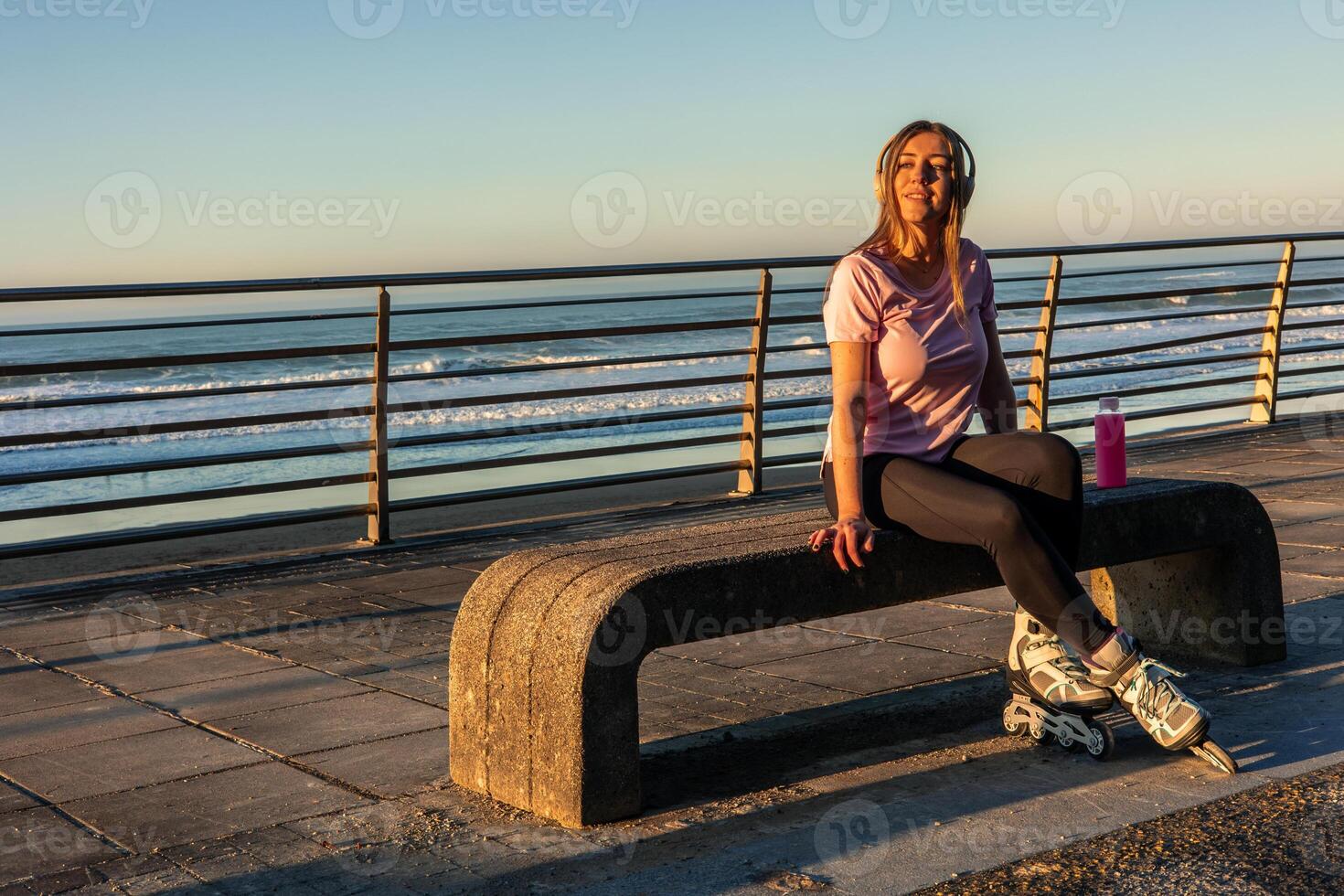 Sportswoman resting on bench wearing inline skates listening to music at sunrise seafront promenade photo