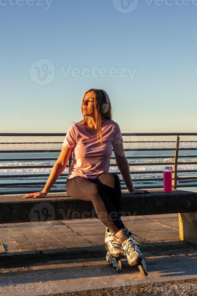 Sportswoman relaxing on bench wearing inline skates and listening music at sunset photo