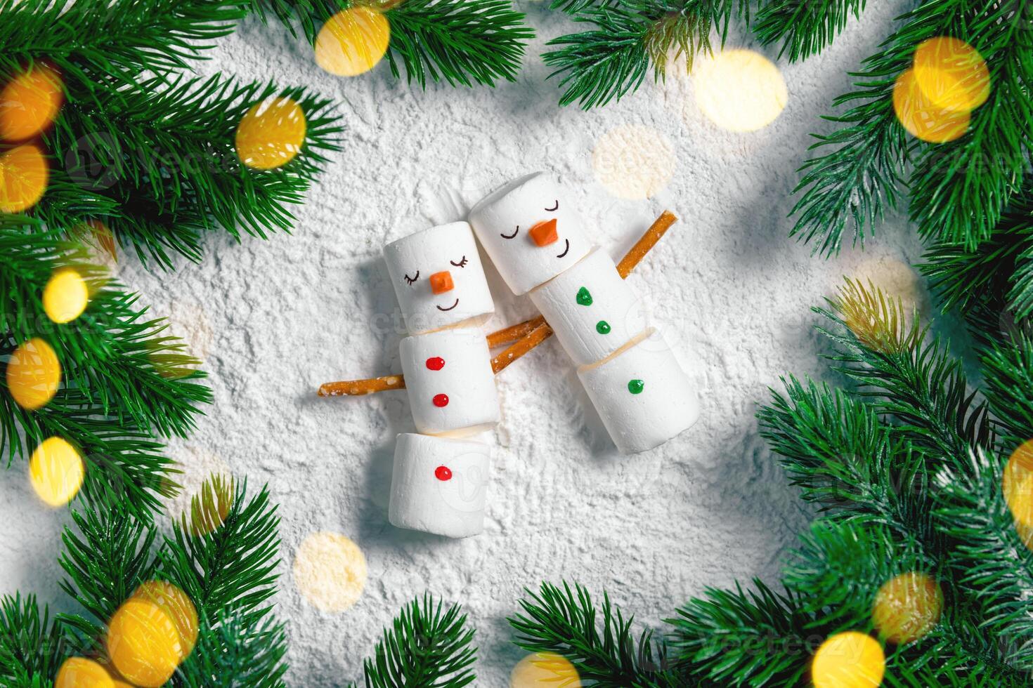 Two adorable marshmallow snowmen are embracing on a snowy background surrounded by christmas tree branches and warm, inviting lights, creating a festive and heartwarming scene photo