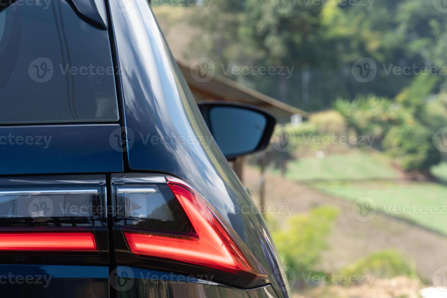 Close-up of a dark blue car tail light and side mirror. with a blurred background of green foliage and wood building in rural areas. photo