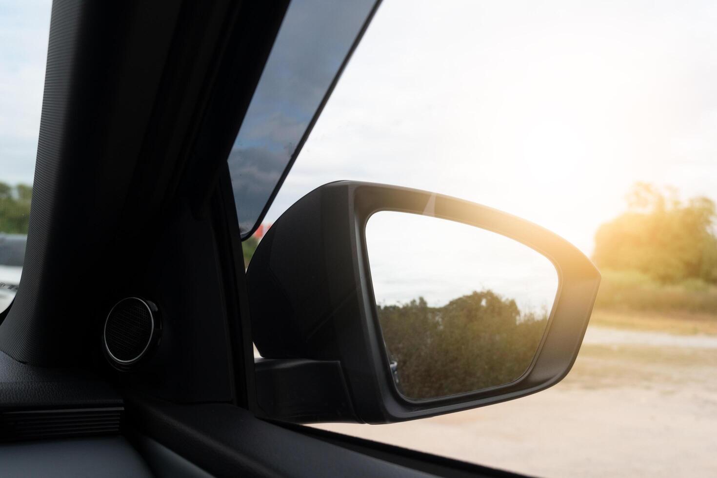 Car side view mirror reflects a blurred landscape and bright sunlight. The interior of the car is visible in the foreground. photo