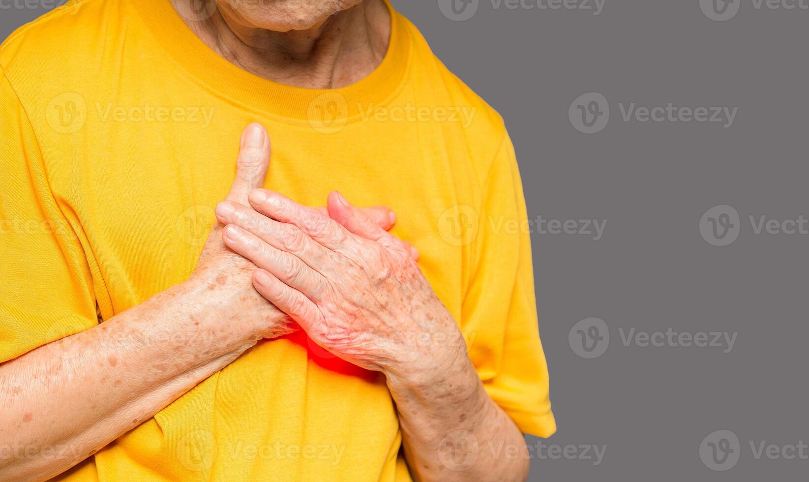 A Senior woman in an orange casual has sore left chest pain while standing on gray background photo