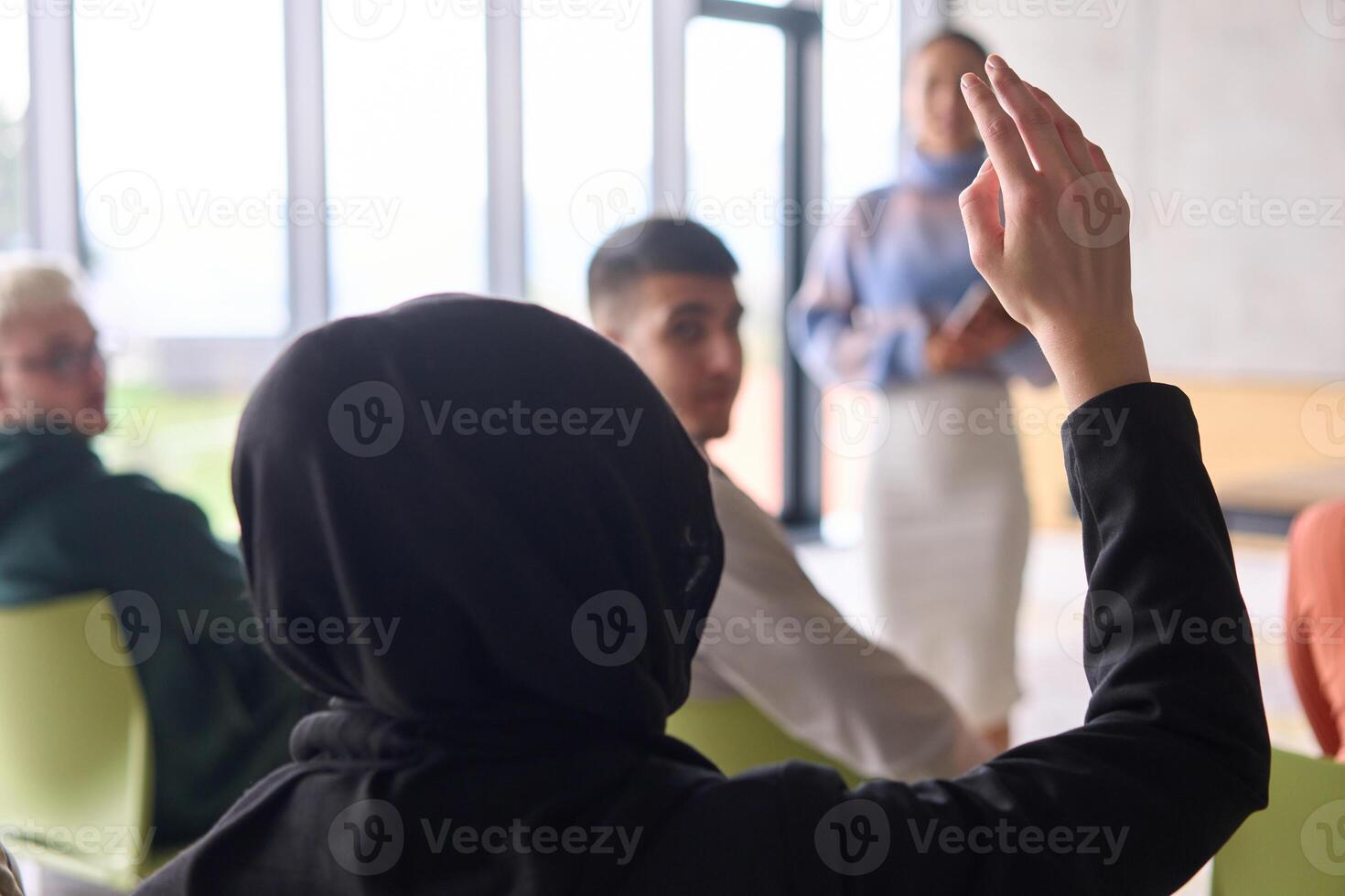 A Muslim businesswoman, dressed in professional attire and wearing a hijab, raises her hand during a business meeting to ask a question to the educator leading the session photo